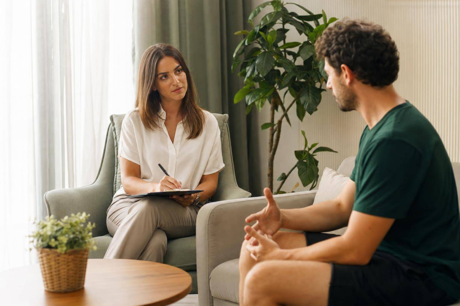 Therapist taking notes while a seated patient describes symptoms
